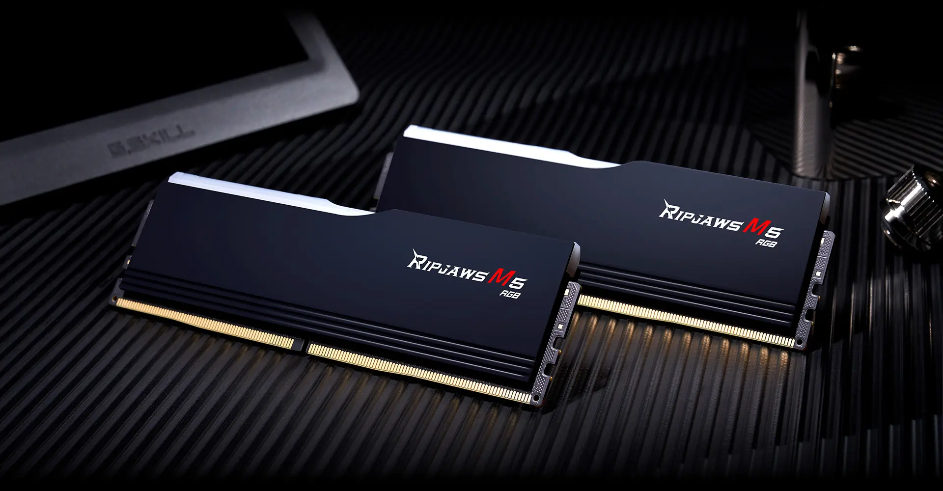 Two matte black G.SKILL Ripjaws M5 RGB DDR5 modules lying on a dark surface