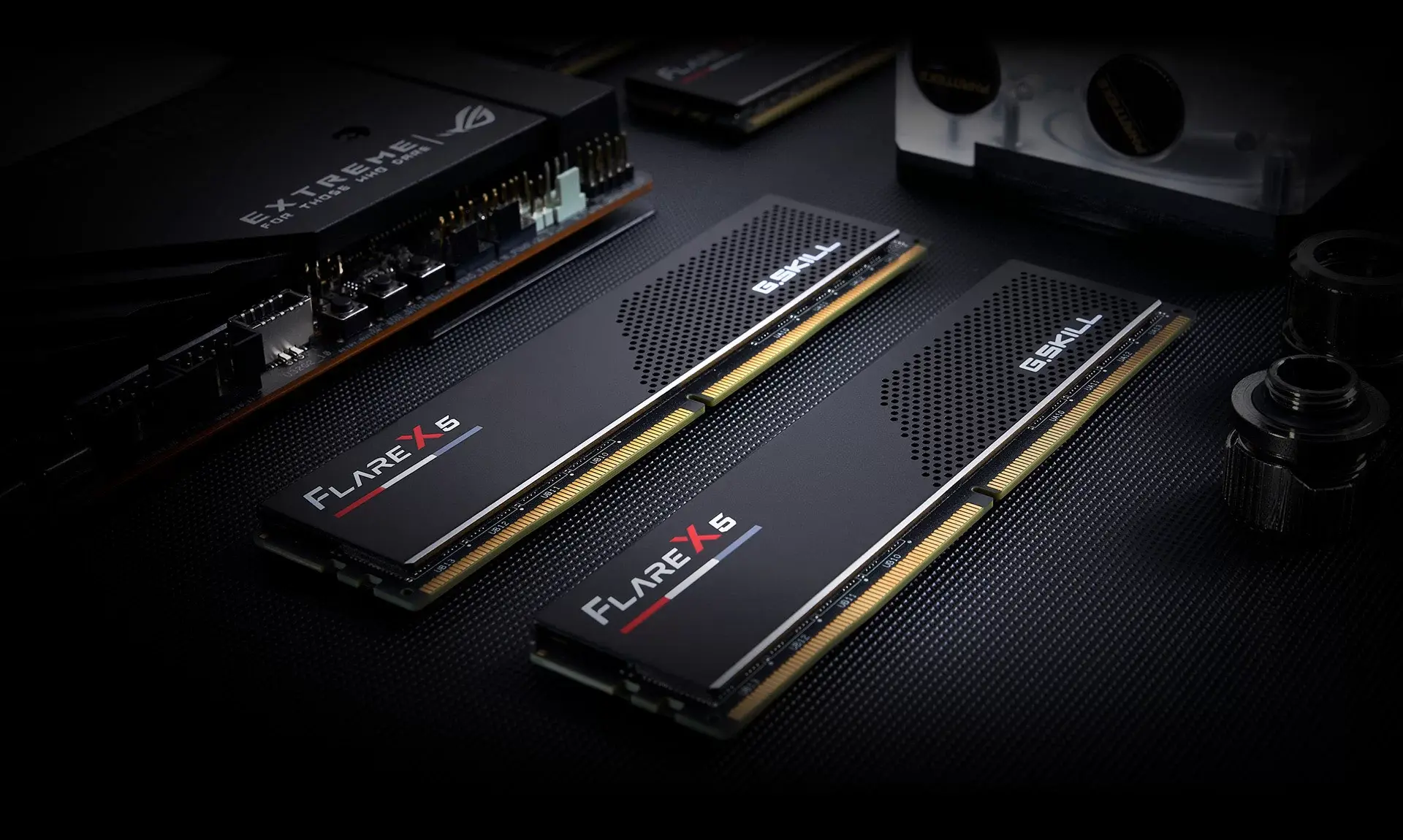 Two matte black G.SKILL Flare X5 DDR5 modules lying on a dark surface beside water-cooling fittings