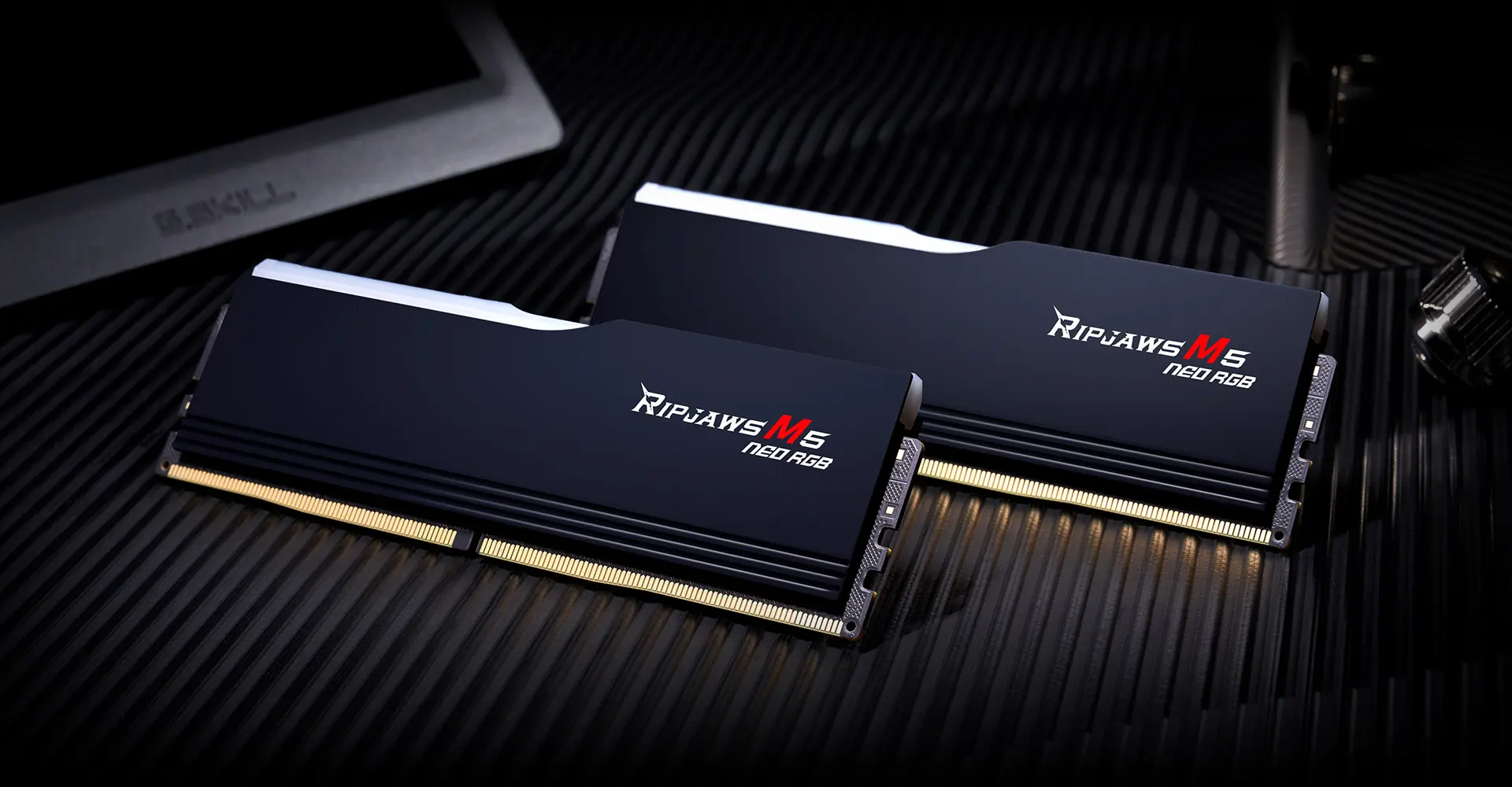 Two matte black G.SKILL Ripjaws M5 Neo RGB DDR5 modules lying on a dark surface